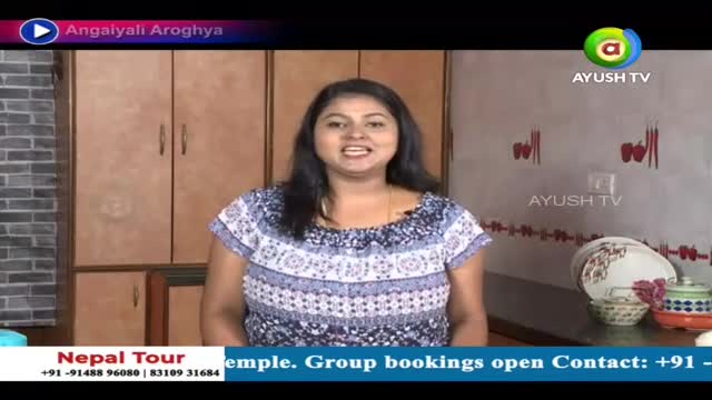 A woman in a patterned top is speaking directly to the camera, likely on a show from Ayush TV. Behind her are wooden cabinets and a display of dishes.
