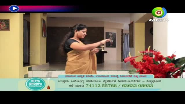 A woman in a gold sari holds something up, possibly a small lamp, as she stands in a doorway.  The Ayush TV logo is visible in the corner, and the scene appears to be set in a home in India.
