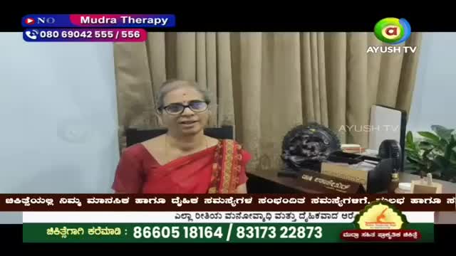 A woman in a red sari speaks from a studio, her glasses glinting. On the desk beside her, a small fan whirs next to a decorative item.