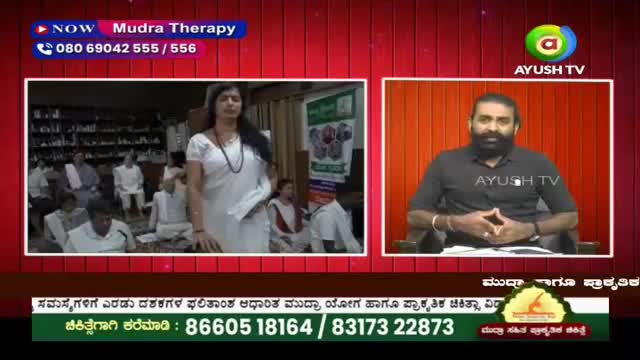 A woman in a white sari stands before an audience, her hands held in a gesture. On a split screen, a man with a beard sits in a chair, his hands clasped.