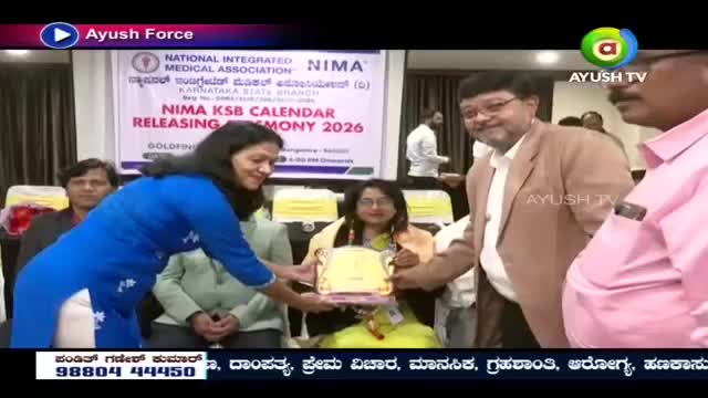 A woman in a bright blue kurta hands a golden plaque to another woman seated in a yellow outfit. A man in a tan jacket stands beside them, smiling as he watches the presentation.