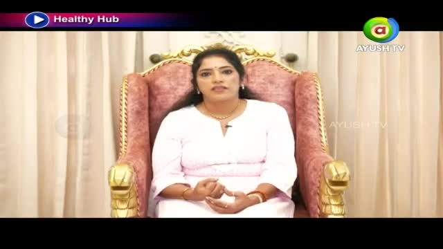 A woman in a white outfit sits in a plush, ornate chair. She speaks directly to the camera, her hands clasped in her lap.