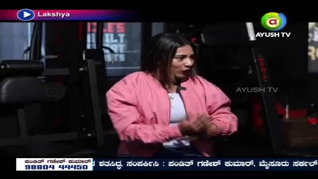 A woman in a pink jacket speaks with her hands clasped, sitting in front of gym equipment. The Ayush TV logo appears on screen.