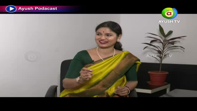 A woman in a bright yellow sari speaks animatedly, her hands gesturing. A potted plant with dark leaves sits nearby on a small table.