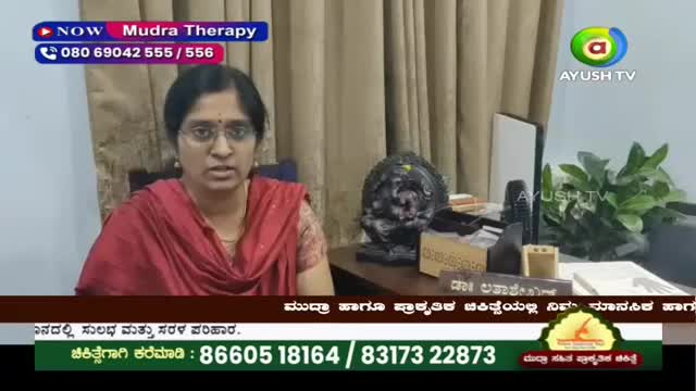 A woman in a red sari speaks directly to the camera, her glasses reflecting the studio lights. Behind her, a dark, ornate sculpture sits on a desk next to a potted plant.