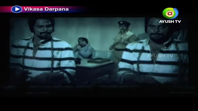 Two men in striped shirts are chained, facing forward. A police officer stands behind a table where another man sits, looking down.