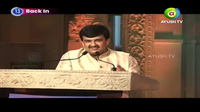 A man in a cream-colored kurta stands at a podium, speaking into two microphones. The Ayush TV logo appears on the right side of the screen.