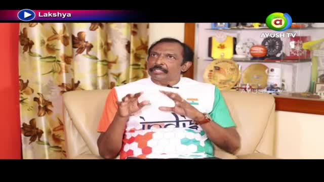 A man in a white shirt with the Indian flag colors speaks animatedly, his hands gesturing. Behind him, shelves display trophies and memorabilia.