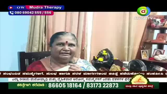 A woman speaks into a microphone, her sari a vibrant splash of color against the muted background. Beside her, a small, dark statue adorned with yellow flowers sits on a table.