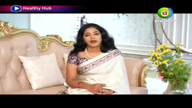 A woman in a white sari speaks from a plush sofa. Yellow flowers in a vase sit to her right.