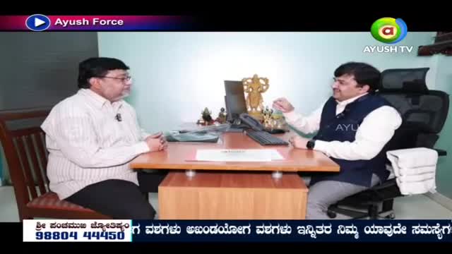 Two men sit across from each other at a desk, engaged in conversation. The man on the right, wearing a dark vest over a white shirt, gestures with his hand as he speaks.