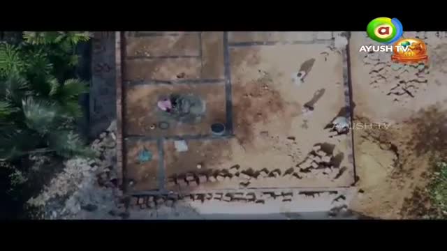 Workers are mixing cement by hand on a construction site in India. Nearby, another person carries a load of bricks.