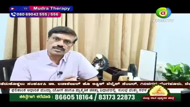 A man with a mustache speaks into a microphone, his light purple shirt buttoned to the collar. Behind him, a beige curtain hangs, and on a desk, a small black fan sits next to a white tablet.