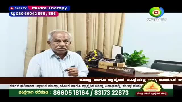A man with gray hair speaks into a microphone, gesturing with his right hand. Behind him, a desk holds a plant and a framed nameplate. The Ayush TV logo is visible in the upper right corner.