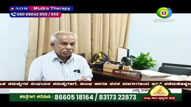 An older man with white hair, wearing a light blue striped shirt, speaks directly to the camera. Behind him, a desk holds a plant and various papers, with a screen displaying "Mudra Therapy" and the Ayush TV logo.
