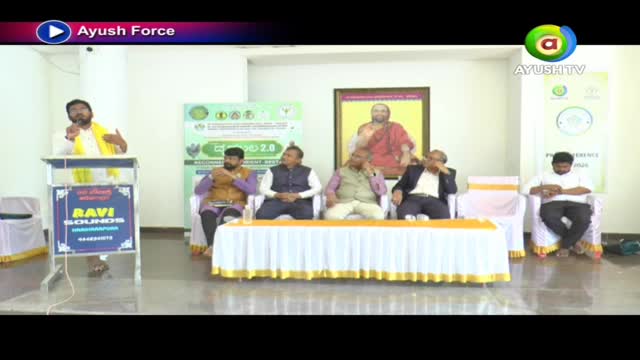 A man in a yellow shawl speaks at a podium, while several men sit on chairs behind a long table. A portrait of a spiritual leader hangs on the wall behind them. A man in a yellow shawl speaks at a podium, while several men sit on chairs behind a long table. A portrait of a spiritual leader hangs on the wall behind them.