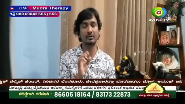 A man in a patterned shirt speaks directly to the camera, his right hand raised in a gesture. Behind him, a shelf holds framed pictures and a dark statue adorned with yellow flowers. A man in a patterned shirt speaks directly to the camera, his right hand raised in a gesture. Behind him, a shelf holds framed pictures and a dark statue adorned with yellow flowers.