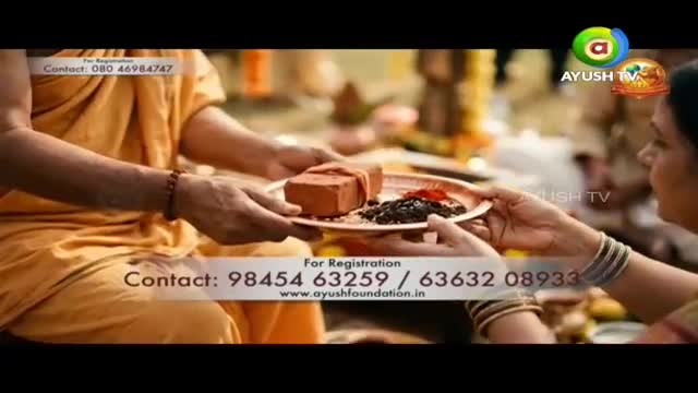 A person in an orange robe offers a plate holding a brick and some dark food. Another person accepts the plate, their bangles glinting. A person in an orange robe offers a plate holding a brick and some dark food. Another person accepts the plate, their bangles glinting.