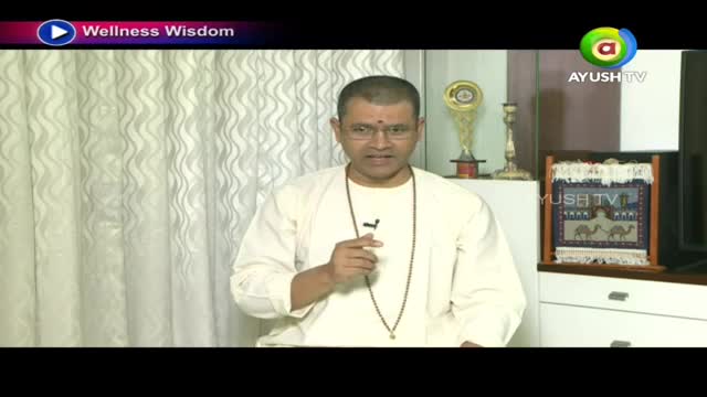 A man in a white kurta speaks into a microphone, gesturing with his right hand. Behind him, a framed tapestry with camels hangs on a shelf beside a trophy. A man in a white kurta speaks into a microphone, gesturing with his right hand. Behind him, a framed tapestry with camels hangs on a shelf beside a trophy.