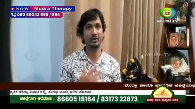 A man with dark hair and a patterned shirt speaks directly to the camera, his left hand gesturing. To his right, a small, dark statue of Ganesha, adorned with yellow flowers, sits on a shelf next to framed pictures. A man with dark hair and a patterned shirt speaks directly to the camera, his left hand gesturing. To his right, a small, dark statue of Ganesha, adorned with yellow flowers, sits on a shelf next to framed pictures.