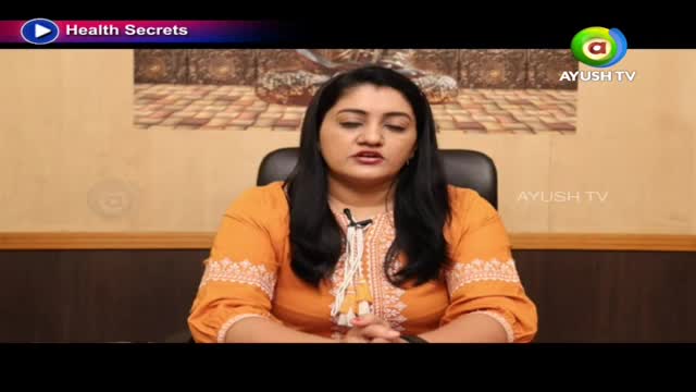 A woman in an orange kurta speaks directly to the camera, her hands clasped in front of her. The Ayush TV logo appears in the corner of the screen.