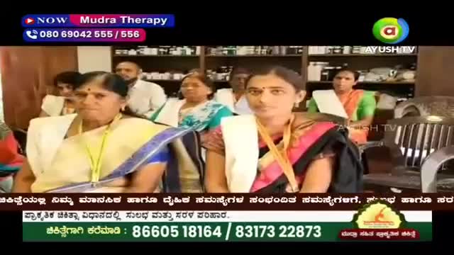 Two women in saris sit attentively, their faces turned towards the camera. Behind them, shelves are packed with bottles, suggesting a pharmacy or clinic.