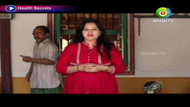 A woman in a bright red outfit stands center, her hands clasped. To her left, a man in a striped shirt and green lungi points with his index finger.
