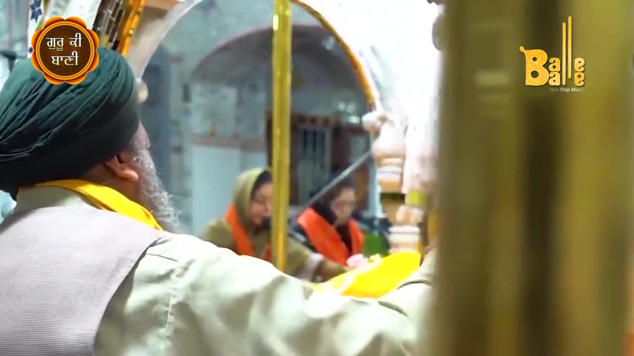 A man in a green turban and a white beard is reaching out, a yellow cloth in his hands. Two women in headscarves sit in the background, their orange scarves bright against the muted interior.
