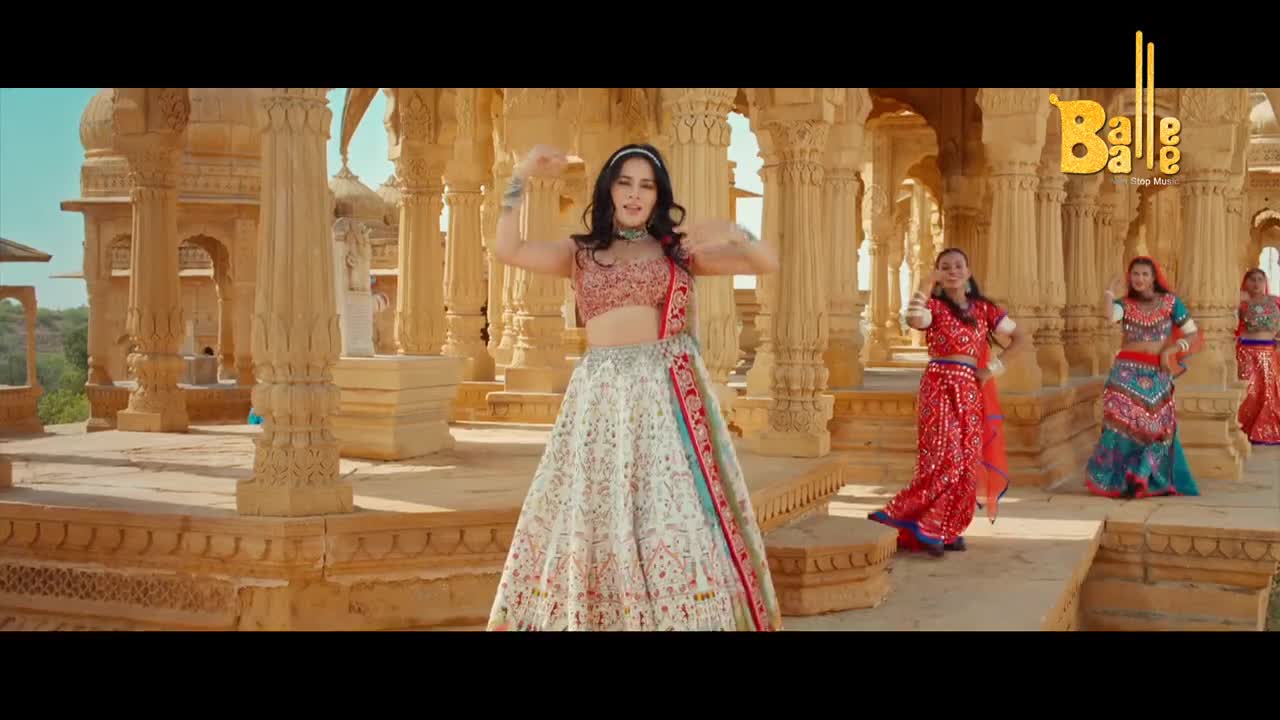In a sun-drenched courtyard, a woman in a jeweled top and flowing skirt dances, her arms raised. Other women in vibrant outfits join her, moving rhythmically to the music of a Balle Balle song.

