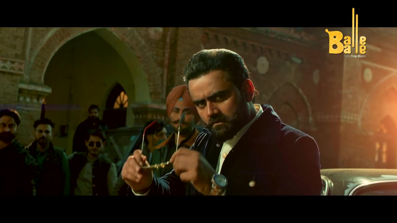 A man in a dark suit holds up his sunglasses, looking directly at the camera. Behind him, a group of men stand in the shadows, and the "Balle Balle" logo is visible in the corner.
