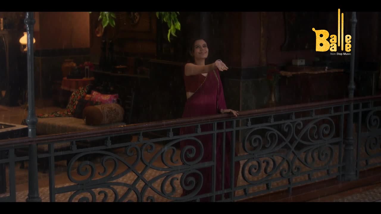 A woman in a maroon sari stands on a balcony, smiling and gesturing with her hand. Rain falls lightly around her, and the Balle Balle logo is visible in the background.
