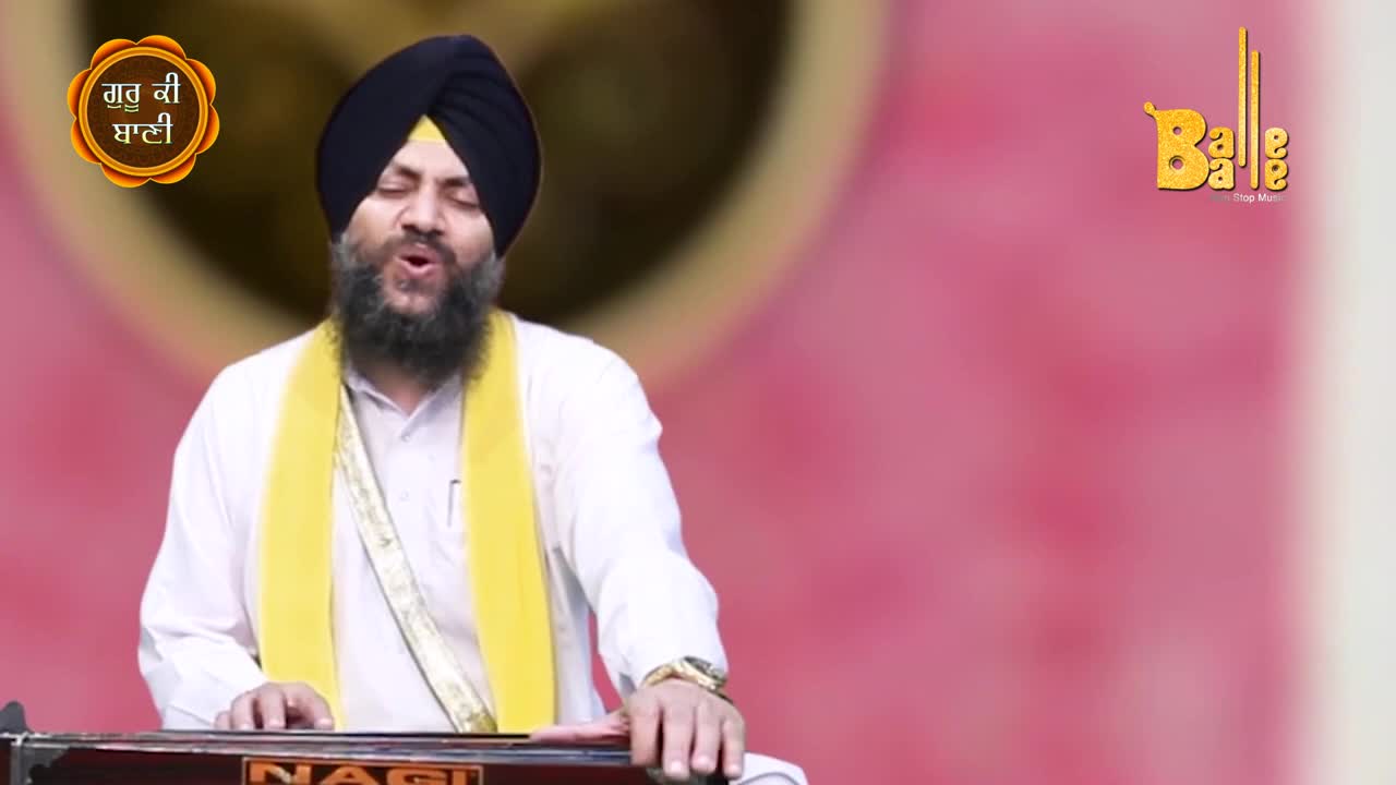 A man in a turban and yellow scarf sings, his voice open and clear. He's playing a harmonium, the keys moving under his hands as he performs for the camera.

