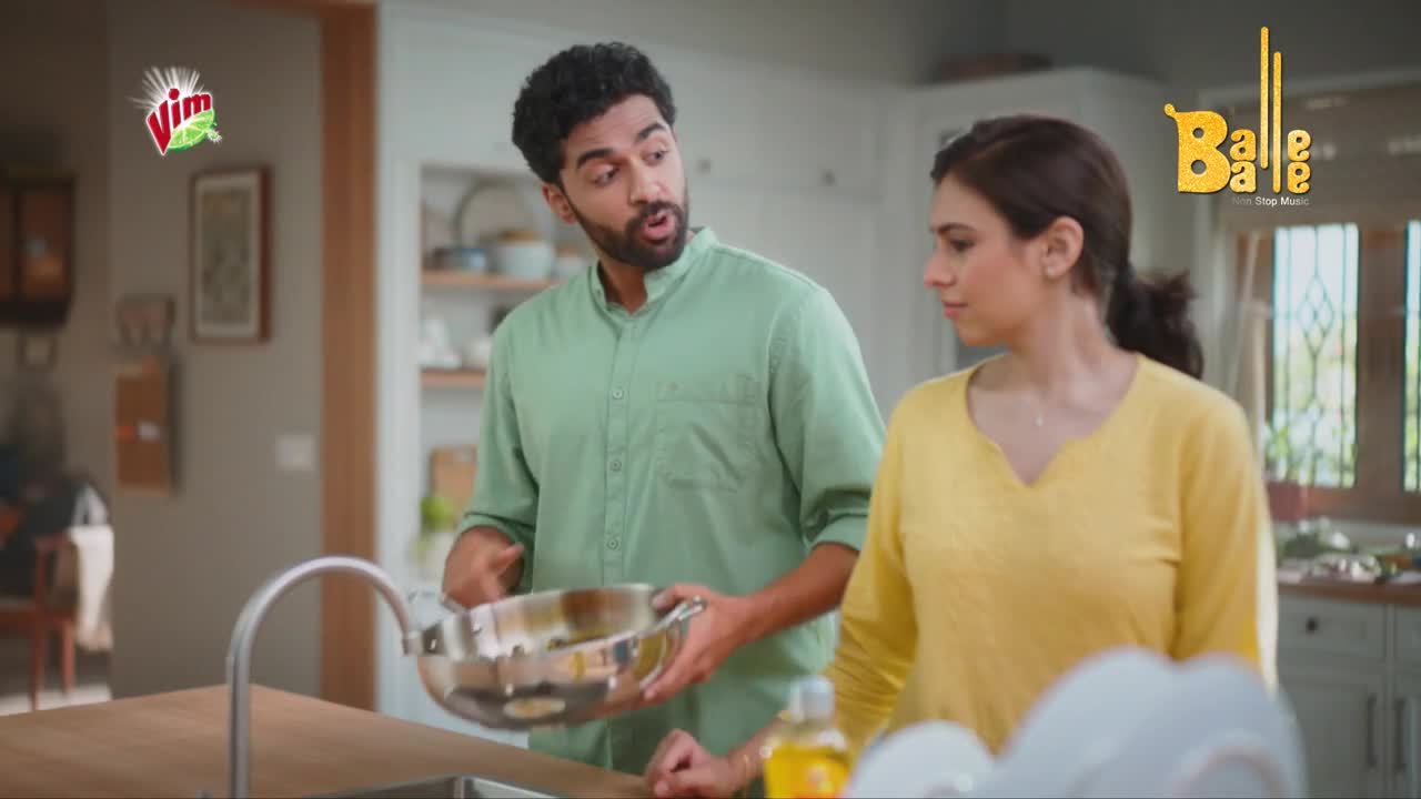 A man, holding a steel pot, gestures emphatically while speaking to a woman in a yellow top. The woman looks on, seemingly listening, as the man appears to explain something near the kitchen sink.
