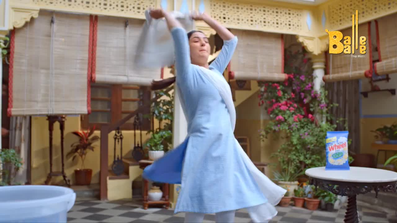 A woman in a light blue salwar kameez twirls, holding a white cloth above her head. The courtyard is bright, and a package of Wheel detergent sits on a small table.
