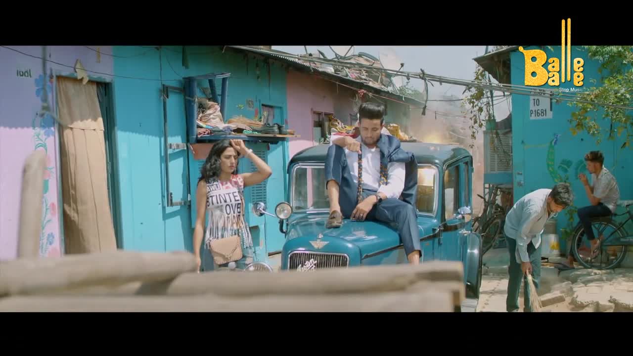 A man in a suit sits on the hood of a vintage blue car, while a woman in a tank top stands nearby, hand on her head. A few other people are milling about on the street, as if this is a scene from a Bollywood music video.
