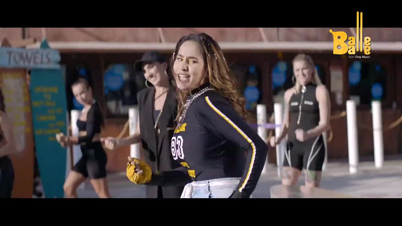 The woman in the center, wearing a black top with yellow stripes and the number 83, is singing and dancing. She's surrounded by others, also moving, in what looks like a music video set.
