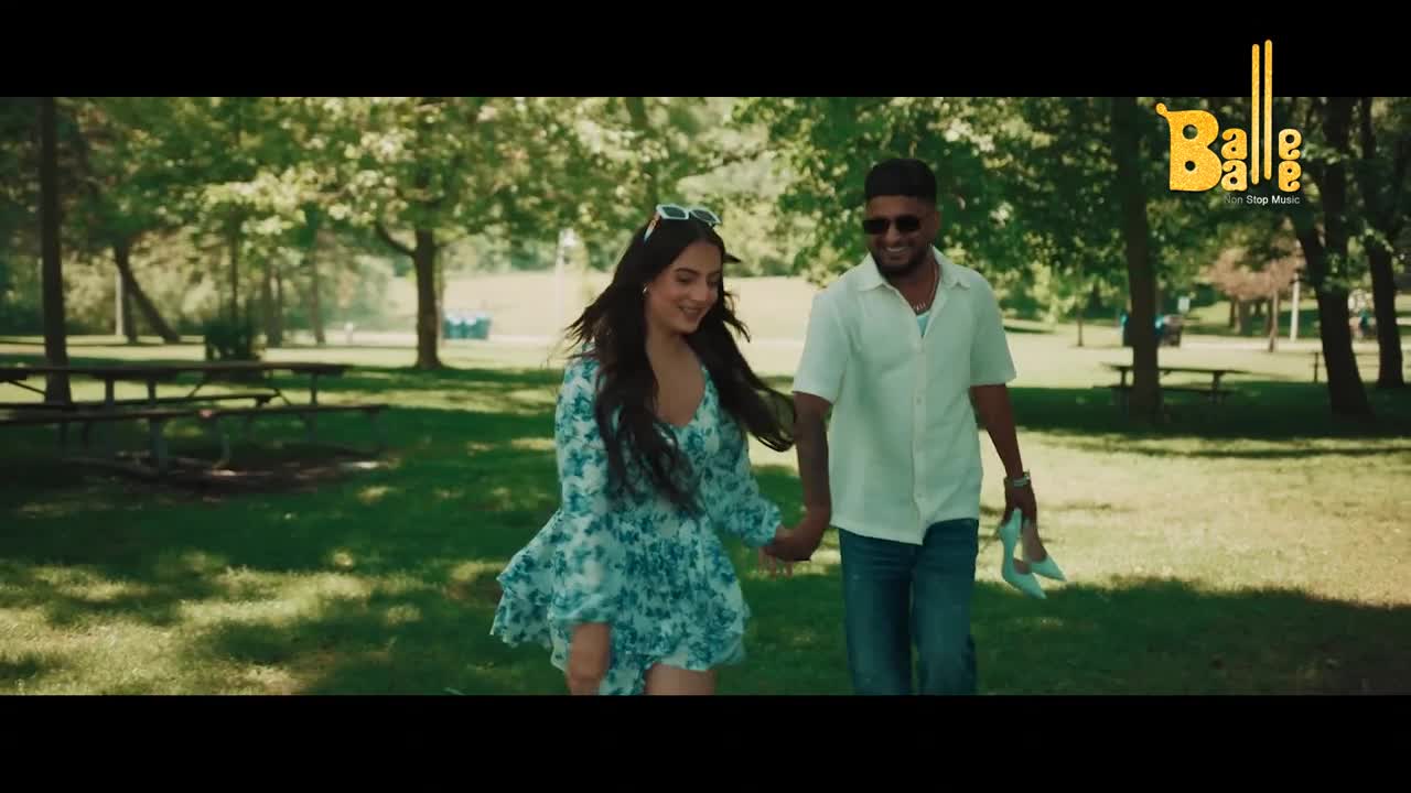 A couple strolls through a park, hand in hand, the woman's dress swirling as they move. The man carries her shoes, and the Balle Balle logo is visible in the corner.
