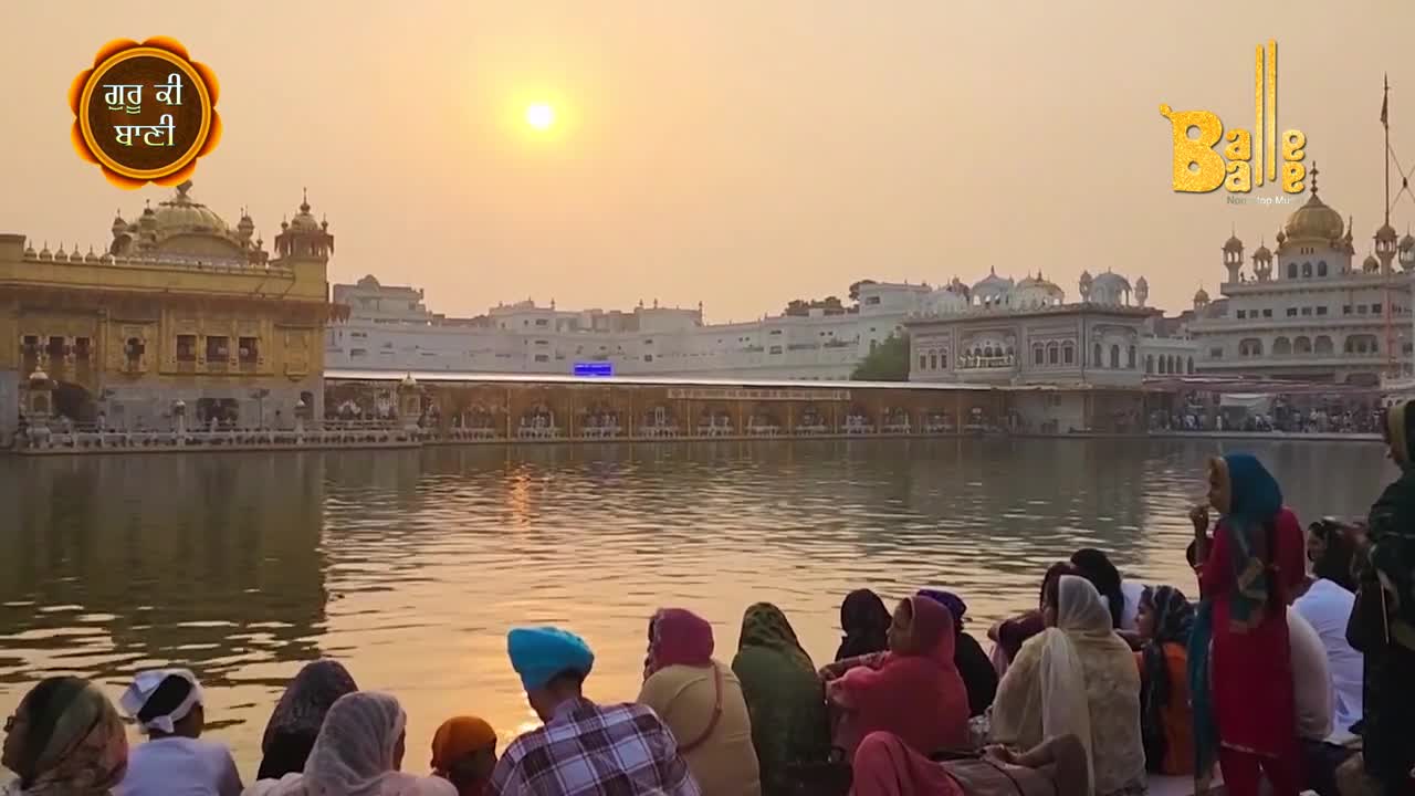 People are gathered near the water, watching the sun's reflection on the golden temple. The sky is a warm, hazy orange, and the water is still. 
