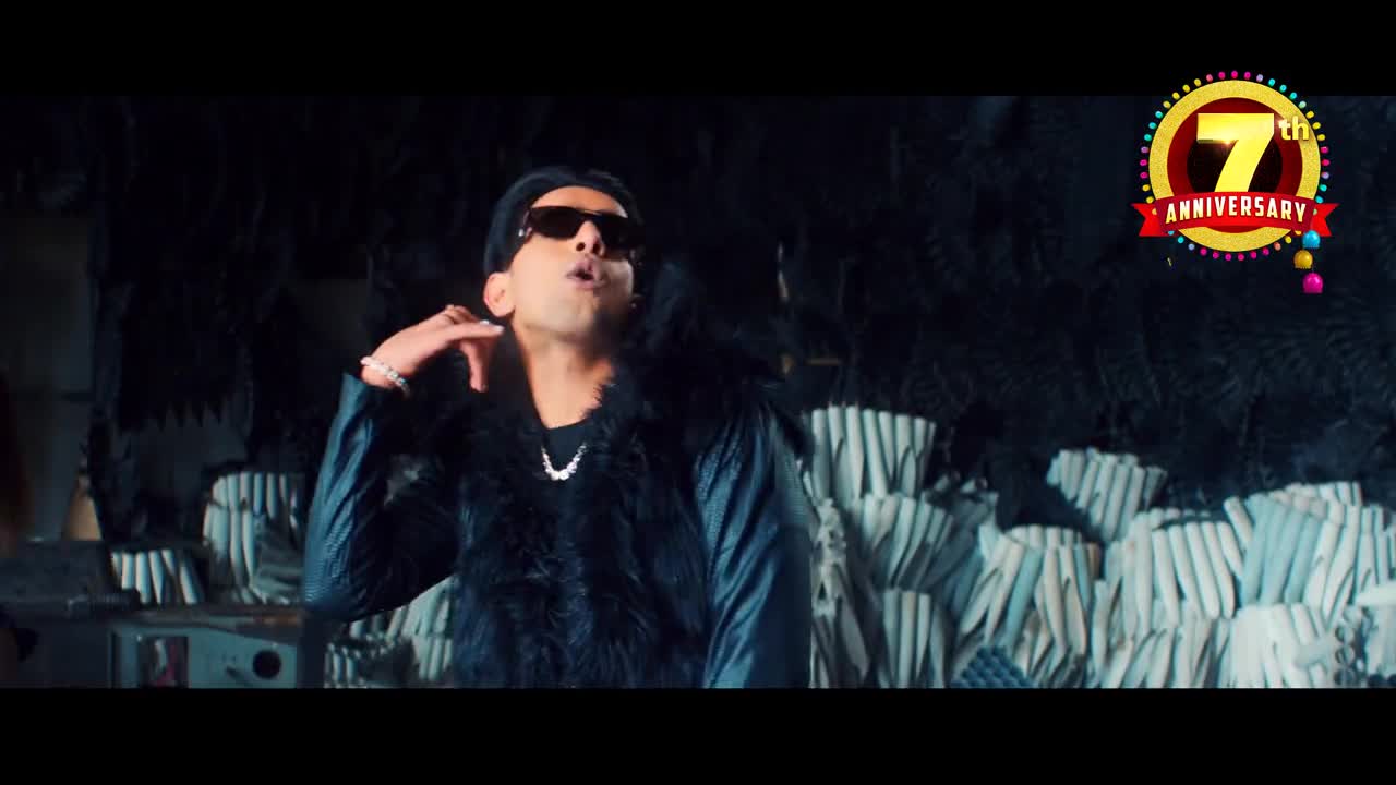 A man in sunglasses and a fur vest is rapping, gesturing with his hand. Behind him, a "7th Anniversary" logo shines, adding to the celebratory vibe.
