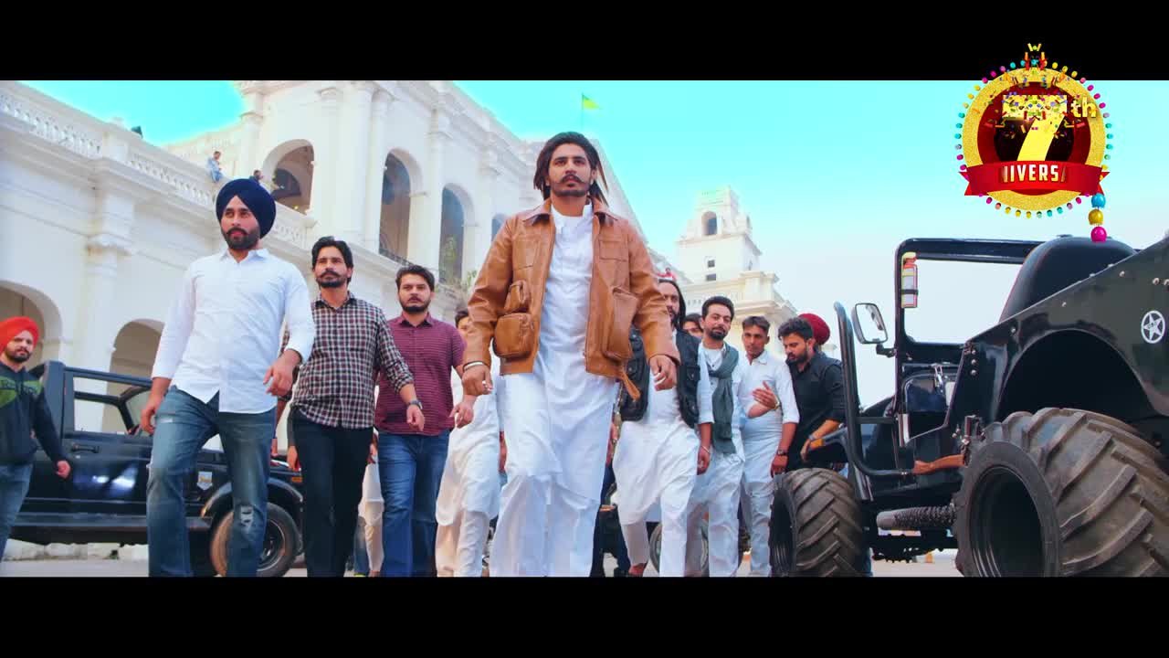 A man in a brown jacket and white kurta leads a procession, followed by a group of men in various outfits.  Two jeeps with oversized tires flank the group as they walk toward the camera.
