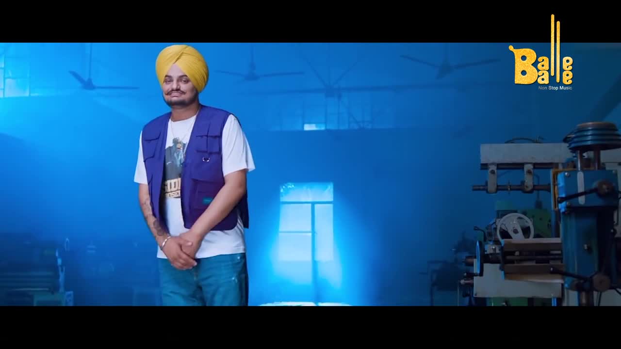 The man in the yellow turban stands in front of a blue backdrop, a factory in the background. He's wearing a blue vest over a white tee, and the "Balle Balle" logo is visible in the corner.
The man in the yellow turban stands in front of a blue backdrop, a factory in the background. He's wearing a blue vest over a white tee, and the "Balle Balle" logo is visible in the corner.
