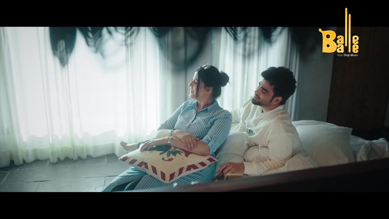 A couple sits on a bed, the woman in a blue striped shirt and the man in white, both looking toward the window. The Balle Balle logo sits in the corner, a hint of the music video's setting.
A couple sits on a bed, the woman in a blue striped shirt and the man in white, both looking toward the window. The Balle Balle logo sits in the corner, a hint of the music video's setting.