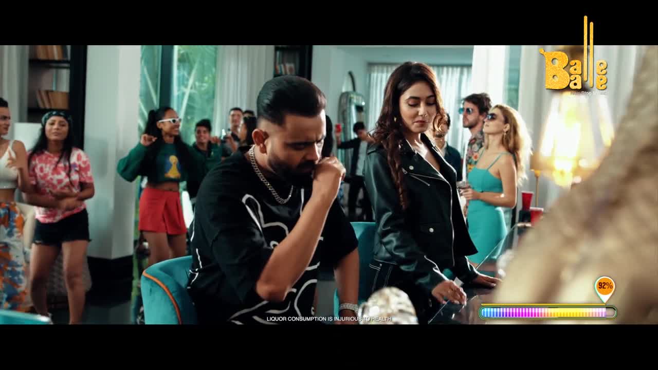 The man in the black t-shirt sits with a pensive look, while the woman beside him stands with a serious expression. A group of people dance in the background, the scene set for a lively Balle Balle music video.
The man in the black t-shirt sits with a pensive look, while the woman beside him stands with a serious expression. A group of people dance in the background, the scene set for a lively Balle Balle music video.