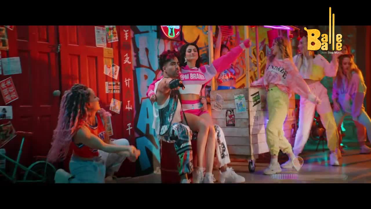A man and woman in matching pink outfits sit on a box, while dancers in colorful tracksuits move around them. The set is vibrant, with graffiti-covered walls and a red door in the background.
