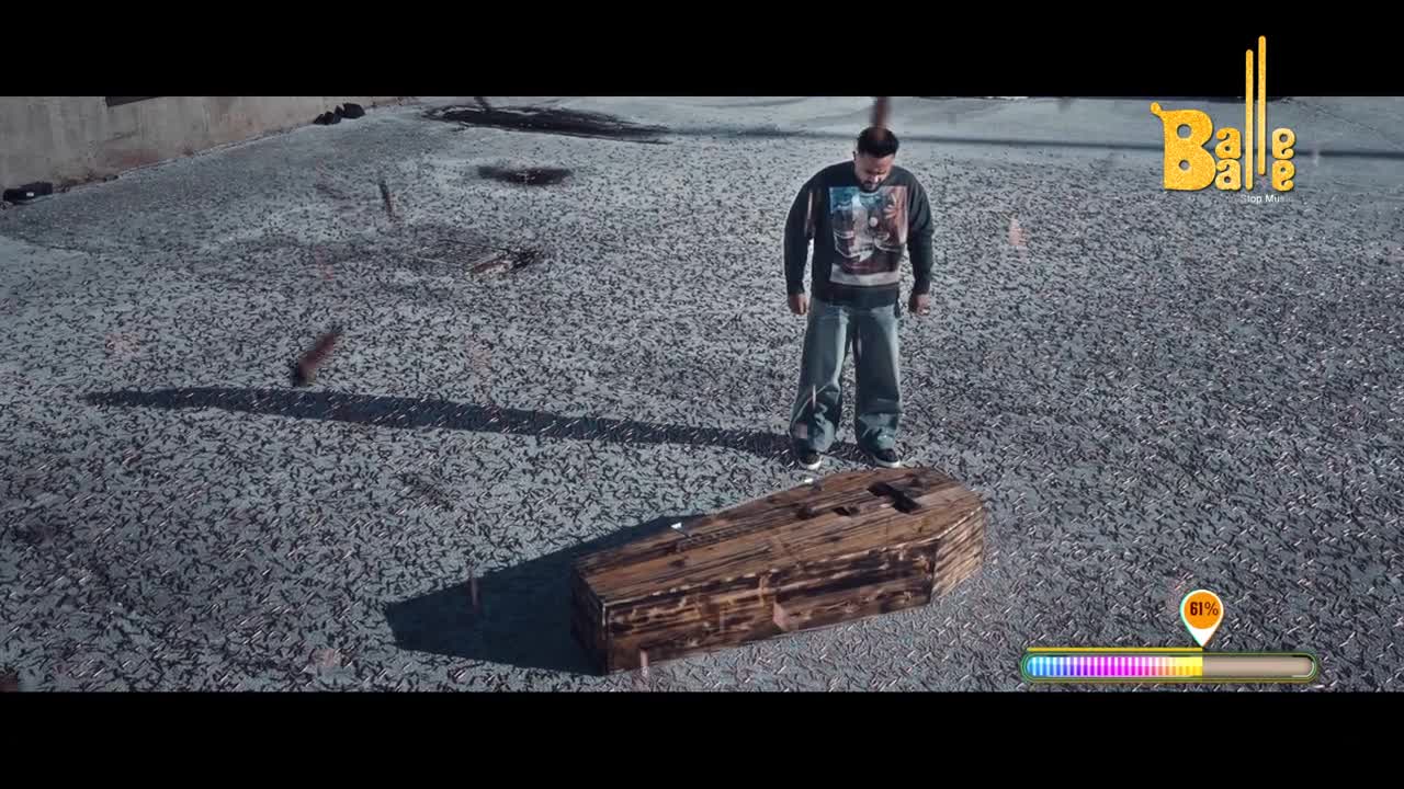 A man in a sweatshirt stands over a wooden coffin on a gravel surface. Small pieces of debris are suspended in the air.

