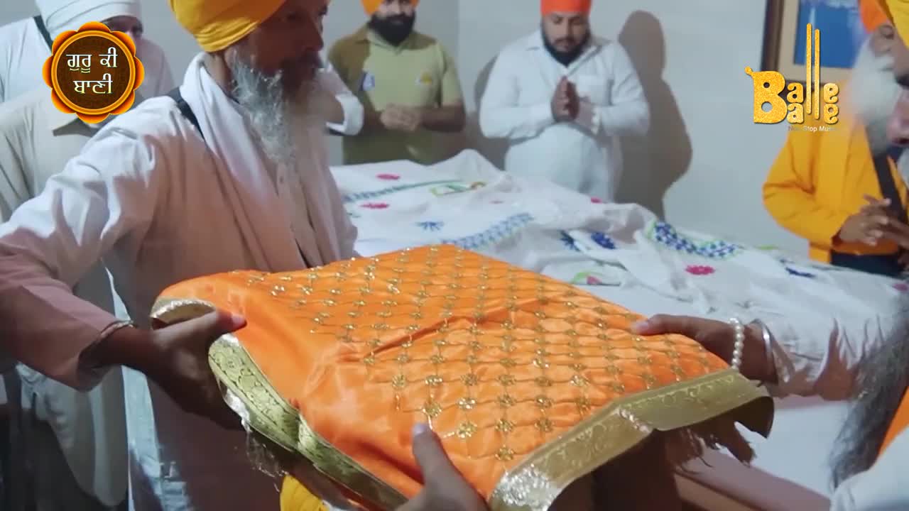 An ornately decorated orange cloth is being carefully presented, held by several hands. Behind, others stand respectfully, hands clasped in prayer.
