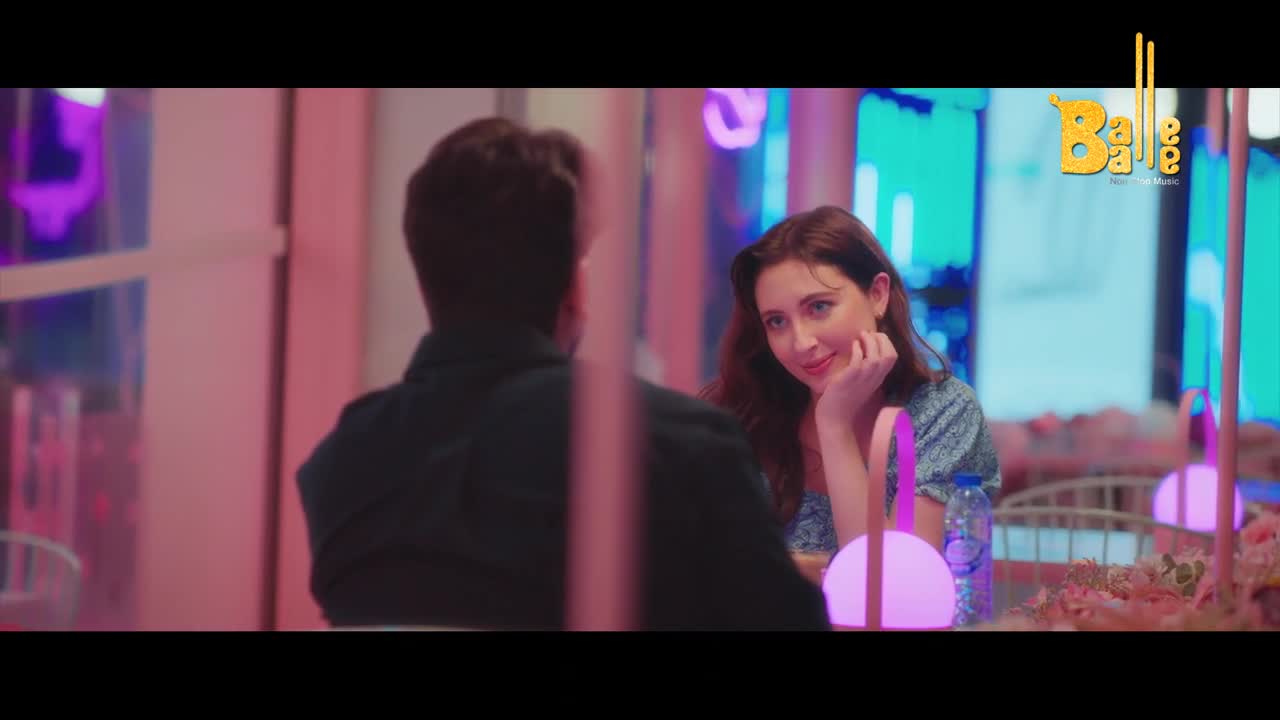 A woman gazes at a man across a table, her hand resting on her chin. The Balle Balle logo appears in the corner, hinting at a Punjabi music video.