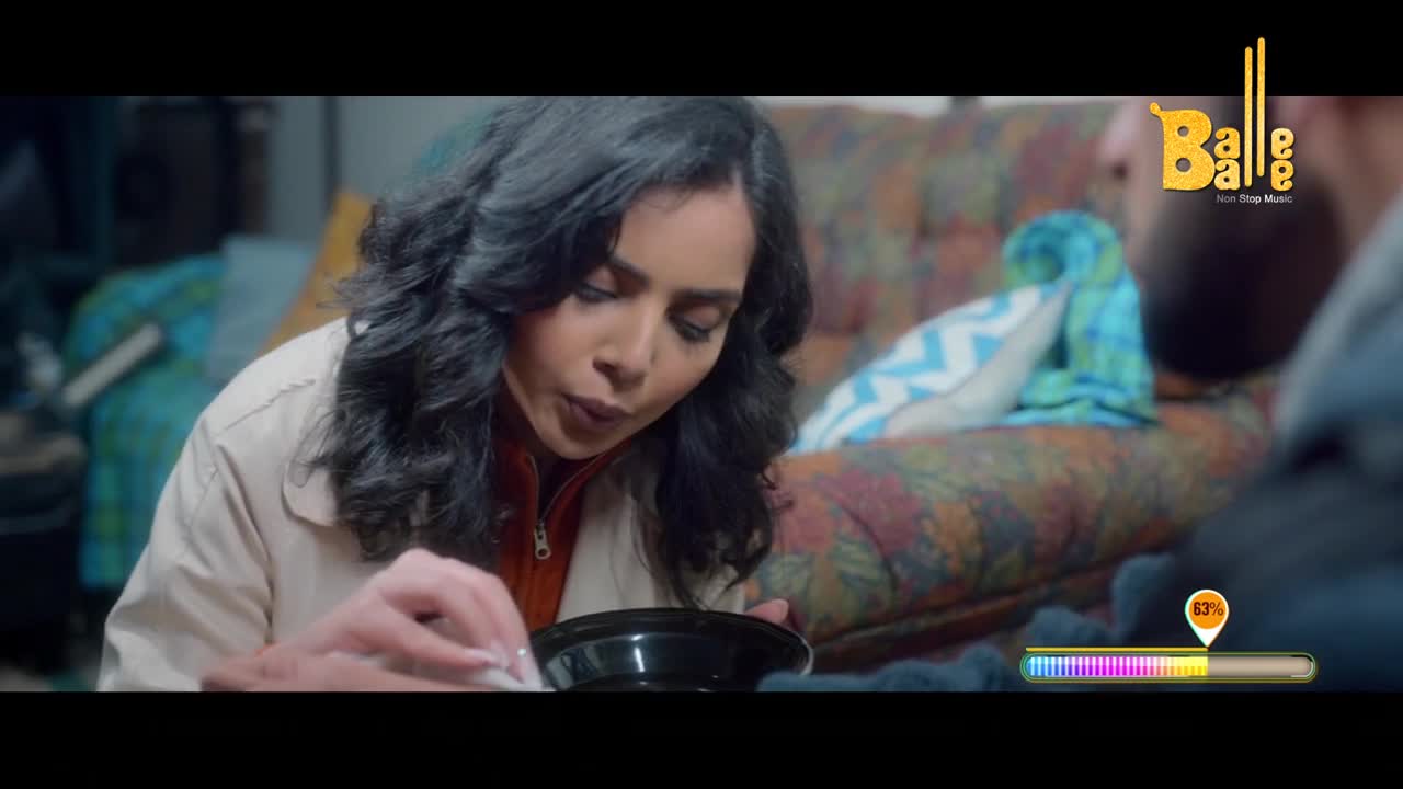A woman with dark, curly hair leans forward, carefully picking something from a black bowl. Beside her, a colorful, patterned sofa is piled with blankets.