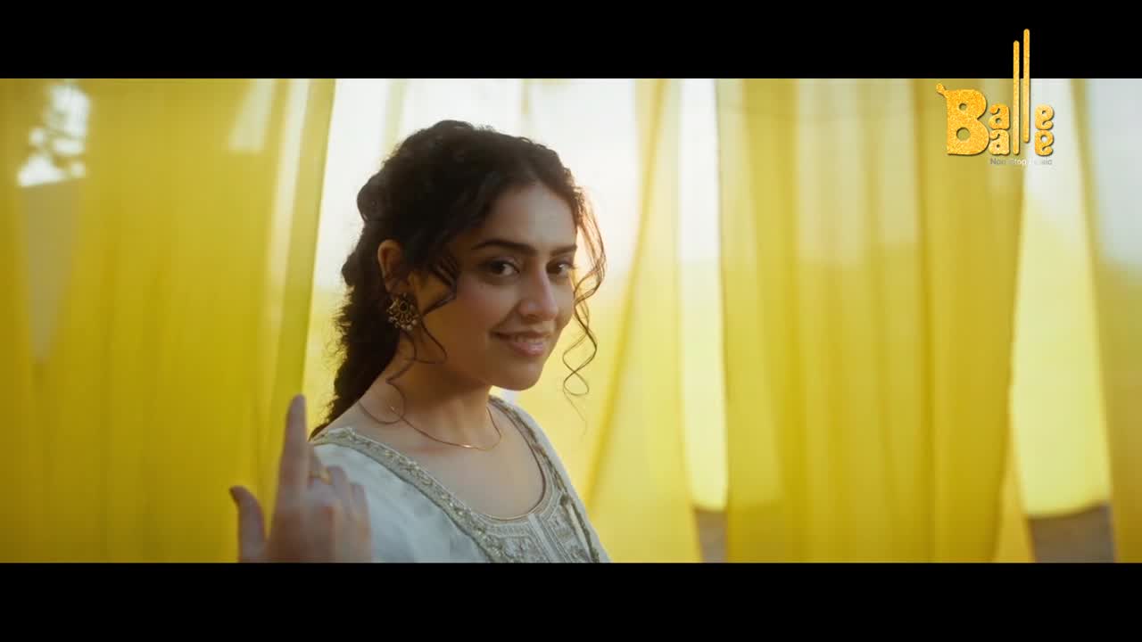 A young woman in a light kurta smiles, her hand raised with a playful gesture. Bright yellow drapes frame her, catching the light.