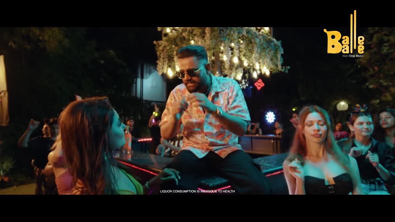 A man in a patterned shirt is perched on a bar, gesturing with his hands as a group of women watch him. Lights twinkle in the background, suggesting a festive night out.